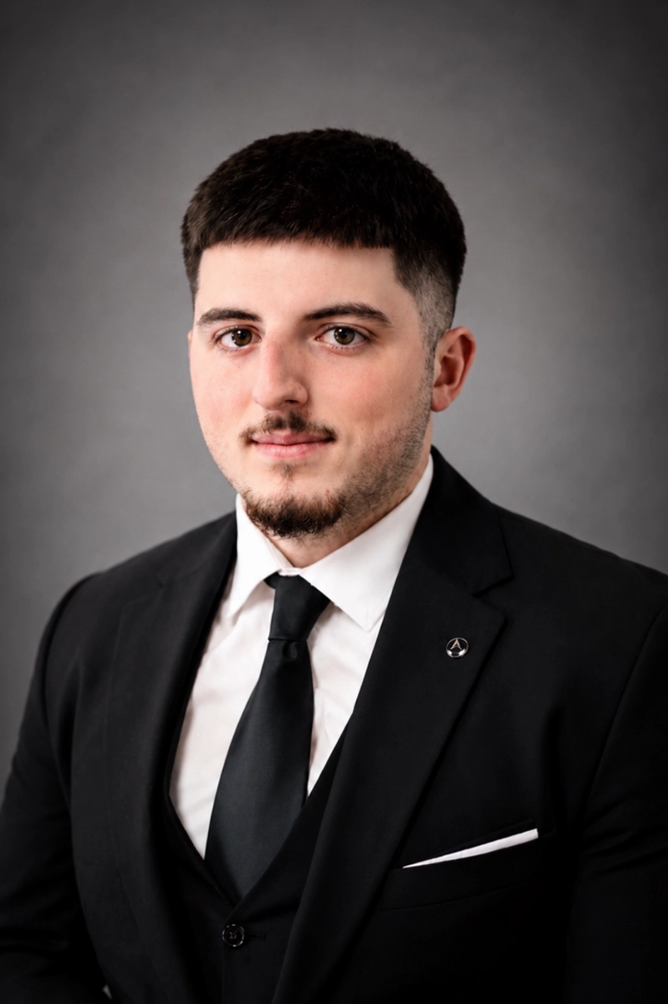 Professional head-and-shoulders portrait of a young man in a black suit, white shirt, and black tie against a gray backdrop.