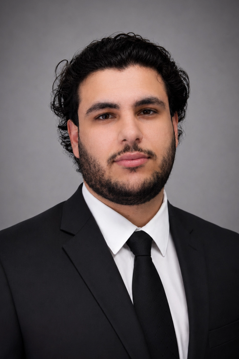 Professional head-and-shoulders portrait of a man with dark hair and beard, wearing a black suit and tie against a gray background.
