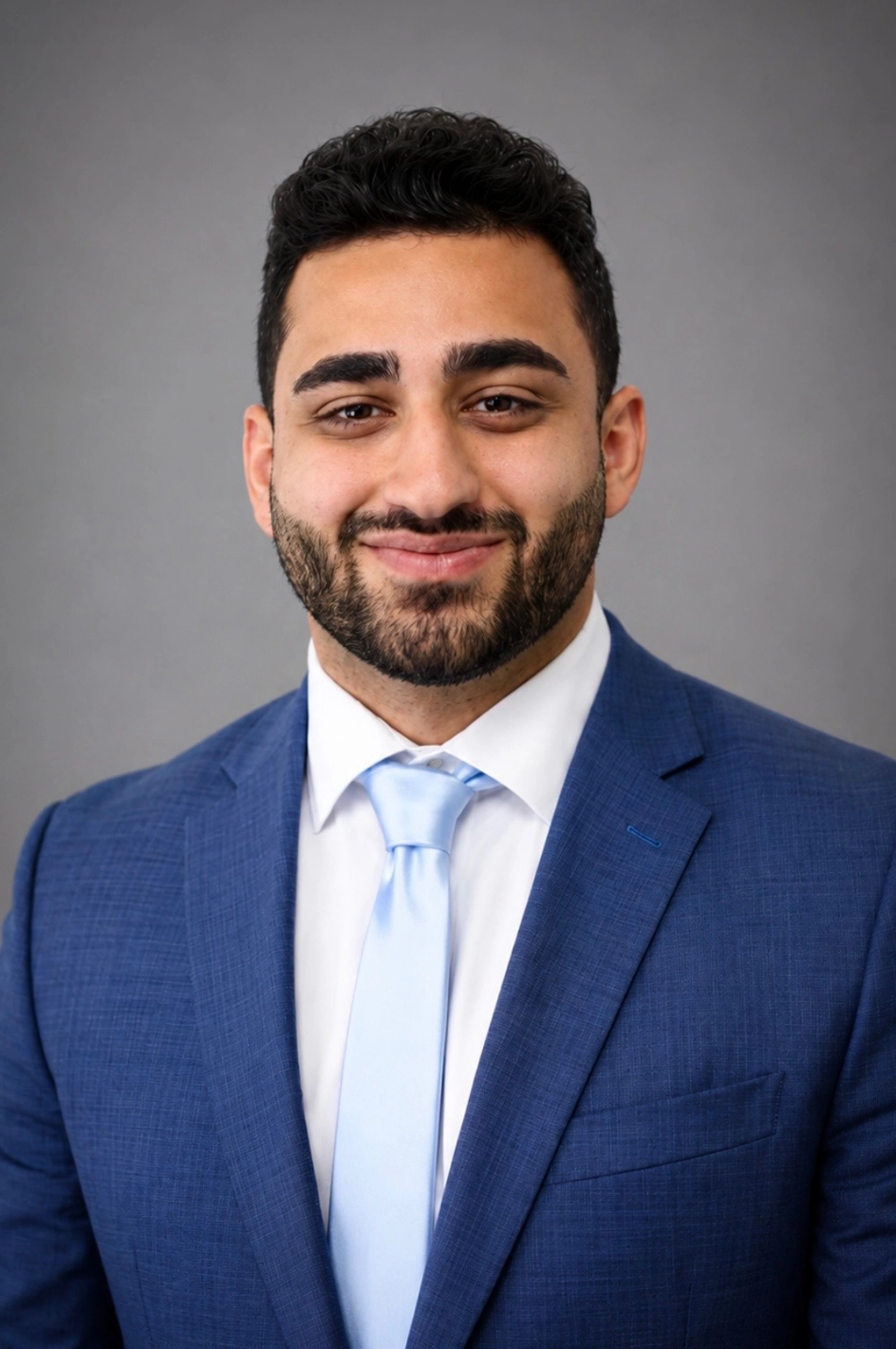 Professional headshot of a man in a blue suit and light blue tie, smiling against a gray background, portrait.