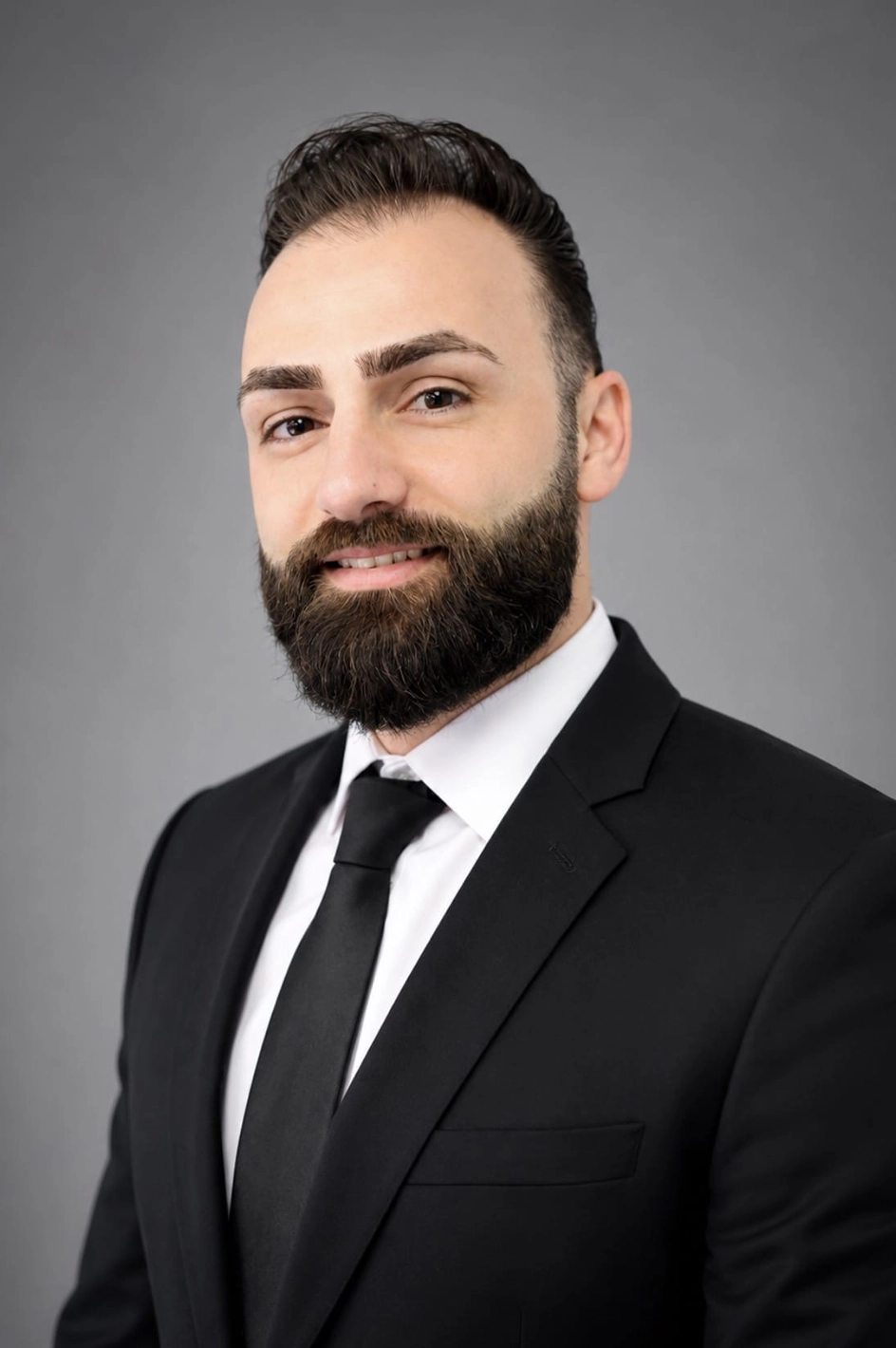 Head-and-shoulders portrait of a man with a full beard in a black suit, white shirt, and black tie, smiling softly against a gray backdrop.
