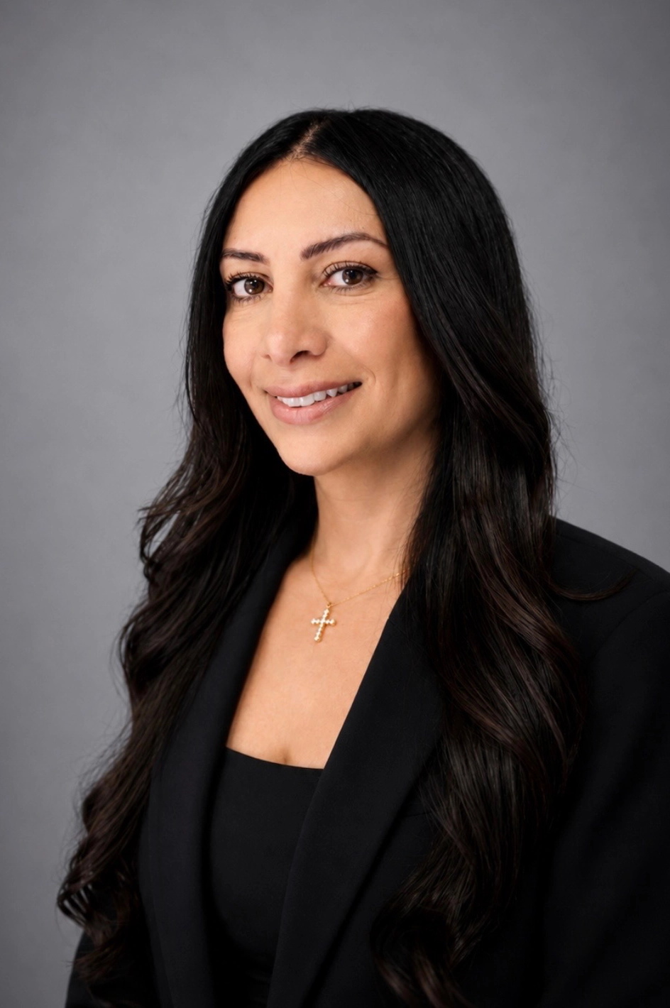 Professional headshot of a smiling woman with long dark hair, wearing a black blazer and a gold cross necklace, against a gray background.